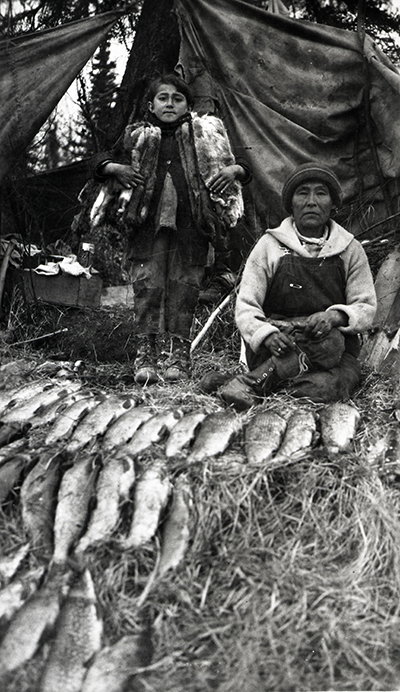Black and white photo of a Dena'ina woman and her daughter with their harvest of fish and pelts circa 1927.