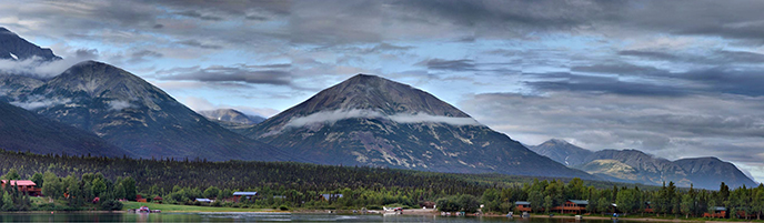 Color photo looking across Hardenburg Bay towards Port Alsworth with Martha's Mountain in the background. Buildings are visible on the shoreline and float-planes in the bay.