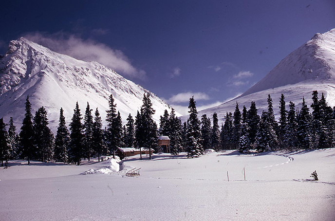 Proenneke stood on frozen Upper Twin Lake and took this photo looking back at the mountains towering over his small cabin in the snow on a sunny day in 1975.
