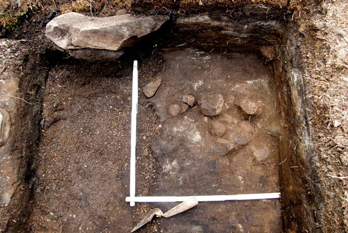 hole dug in the ground with a trowel and ruler in the bottom, near stones