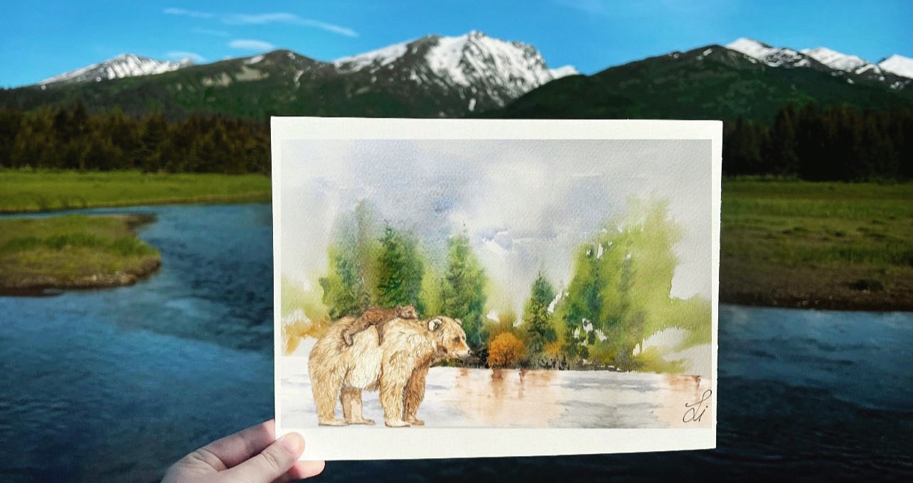 A watercolor painting of a mother bear and cub near a stream, held up in from of a green mountain landscape.