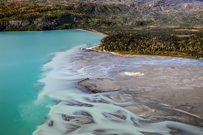 Image of river flowing into a deep blue lake, surrounded by mountains.