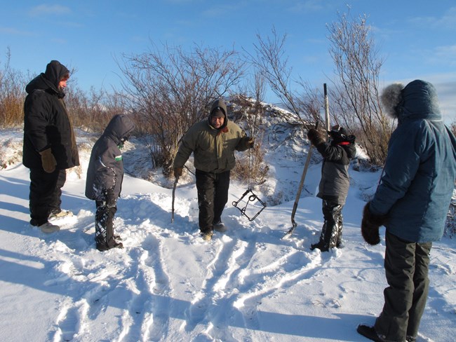 A Dena'ina man holds a metal animal trap in front of a group of people wearing parkas in the snow