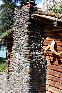 Photo of the backside of a log cabin with a stone chimney and sod roof.