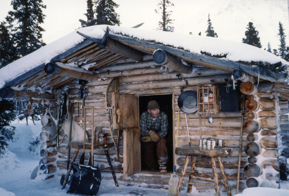 A man wearing a blue flannel shirt and brown pants sits in the open doorway of a log cabin on a winter day.