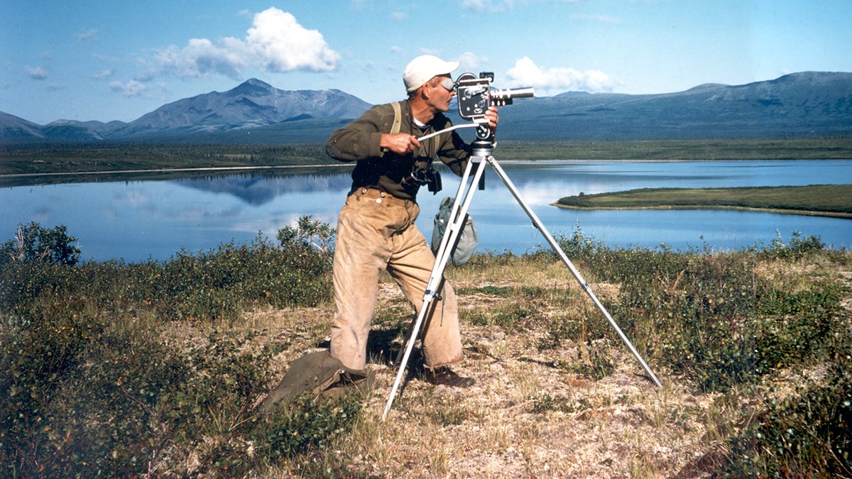 Photo of a man standing in profile filming with an old-fashioned movie camera on a tripod. In the background there is a blue lake and distant mountains.