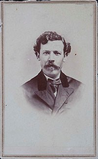 black and white head shot photo of a man with curly hair and a mustache wearing a suit.