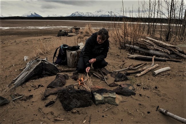 A woman kneeling on a beach builds and lights a fire in a fire ring made of rocks. The beach has large driftwood, tall brown grass, and young trees with no leaves. Snow-covered mountains are in the distance.