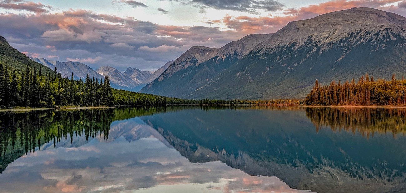 A lake surrounded by dramatic mountains and forest, the sky is dotted with clouds in varying hues. The mountains, trees, and sky are reflected in calm water.