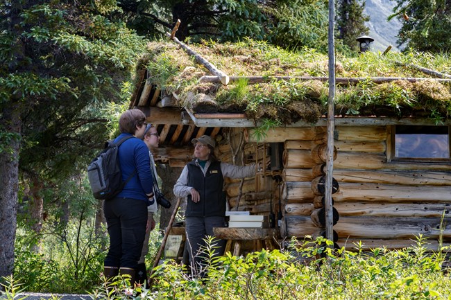 Two women listen to a park volunteer talk about the Richard L. Proenneke cabin. The cabin is surrounded by greenery