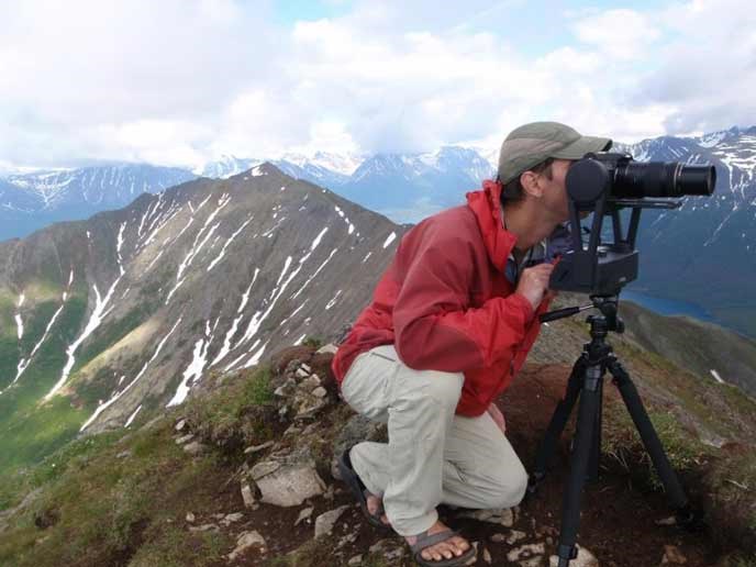 A man shoots photographs from a mountain top with mountains in the background.