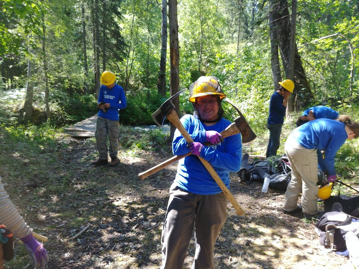 SCA Trail Crew at the Portage Creek Trail a trail crew member wearing a hard hat holds two pulaskis in a cross formation
