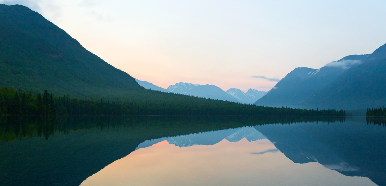 A pink and lavender sunrise over a lake. Mountains with dense green trees are in front of the mountains and the mountains are reflected in the water below.