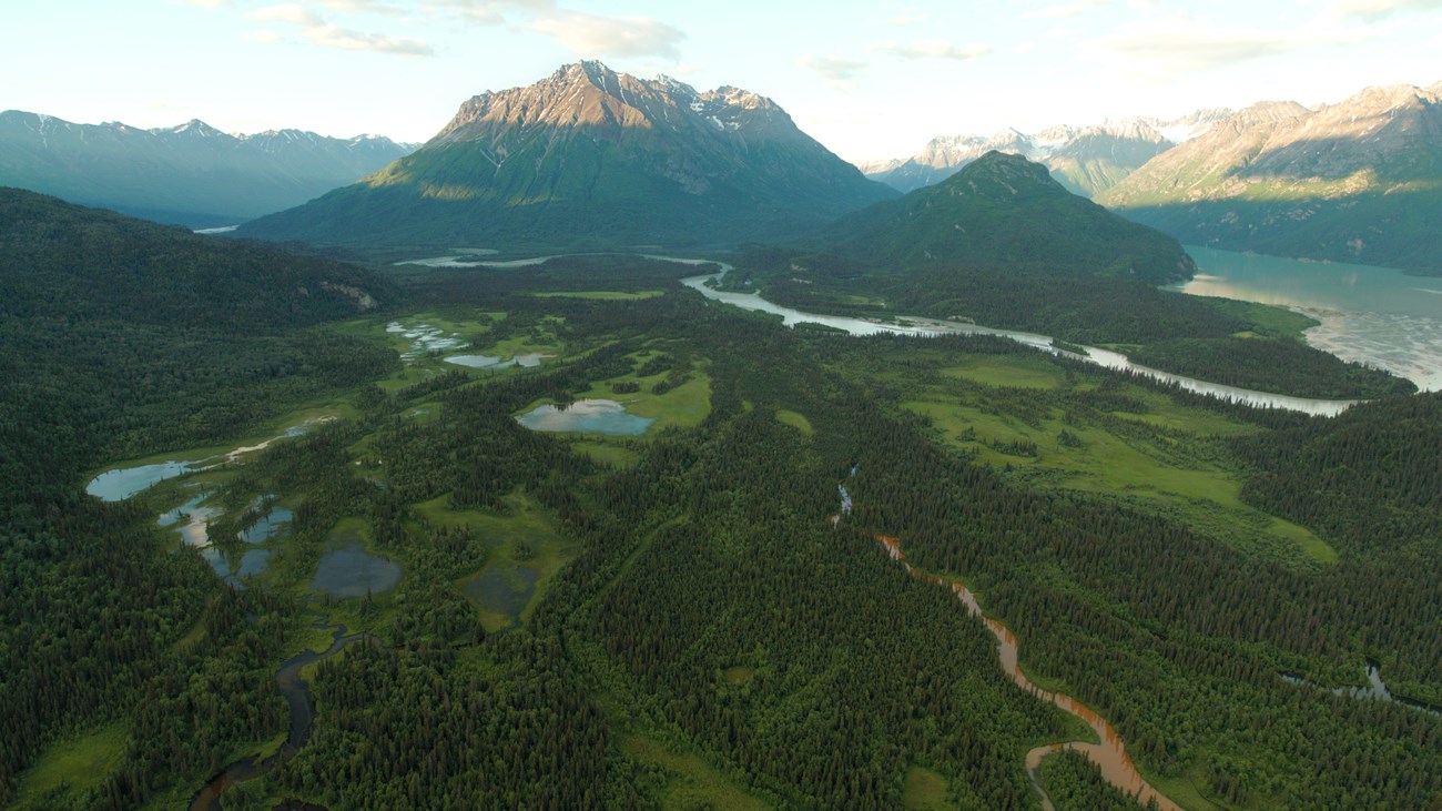 An aerial view of trees, green vegetation, ponds, and winding rivers surrounded by jagged mountains with fading sunlight on the peaks.