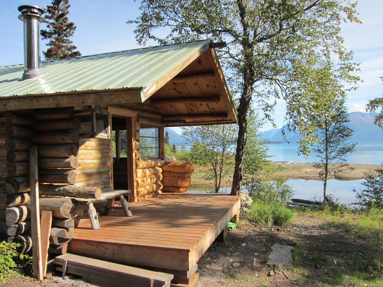 A wooden cabin with a porch is backdropped by water and mountains