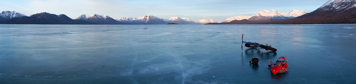 A fat tire bike and backpack rest on the ice of a vast frozen lake bound by snowy mountains. A fat tire bike and backpack rest on the ice of a vast frozen lake bound by snowy mountains.