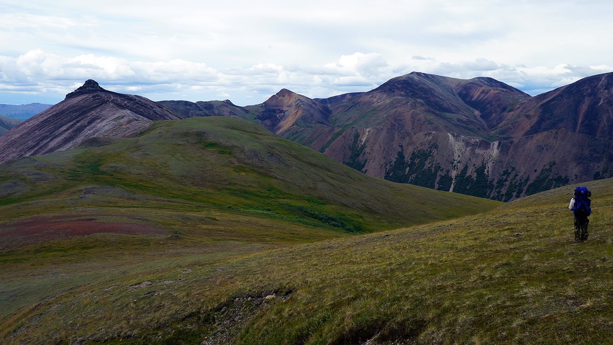 backpacker stands on a tree-less hillside, looking at rolling hills and distant mountains