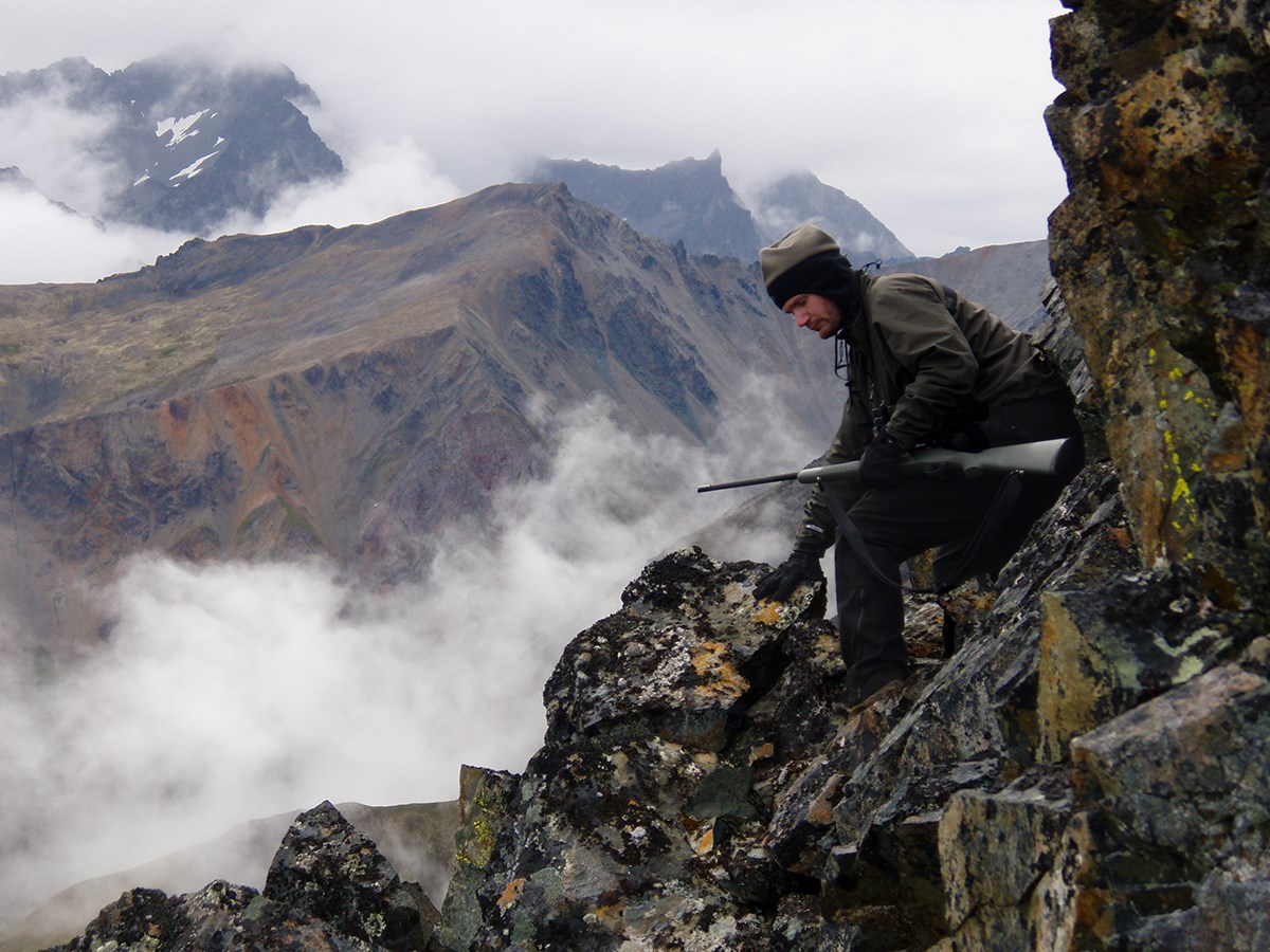man with a rifle navigating steep mountain terrain on a foggy day