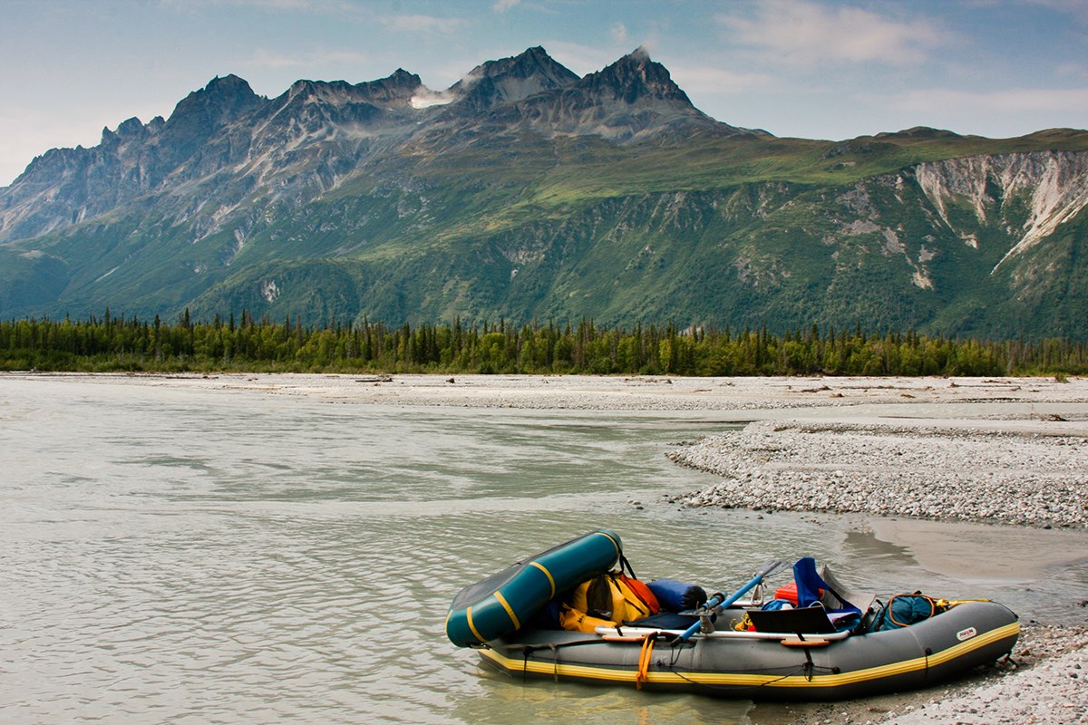 Photo of an inflatable raft loaded with equipment resting on a gravel bar. Tall forested mountains dominate the background.