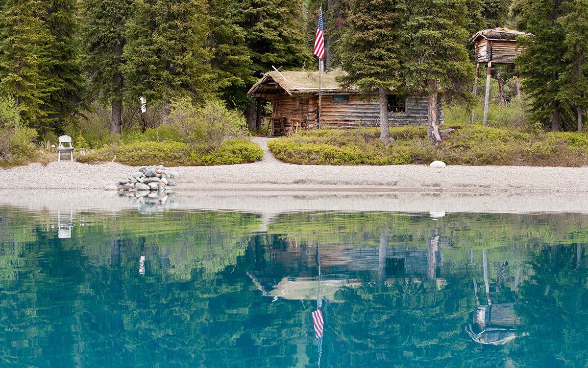 photo of a log cabin tucked into a spruce forest reflecting in a blue lake.