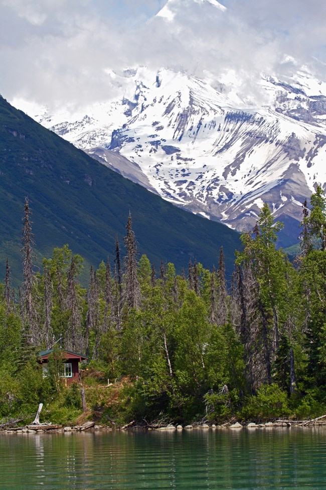 a small cabin on a lake shorewith a snow covered volcano in the background