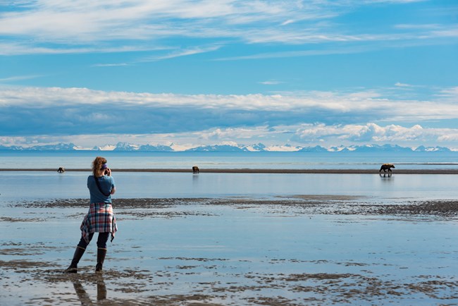 woman standing on mud flat photographing bear in distance.