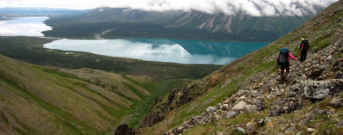 Two people walking on a talus slope above two turquoise hued lakes surrounded by tall mountains.