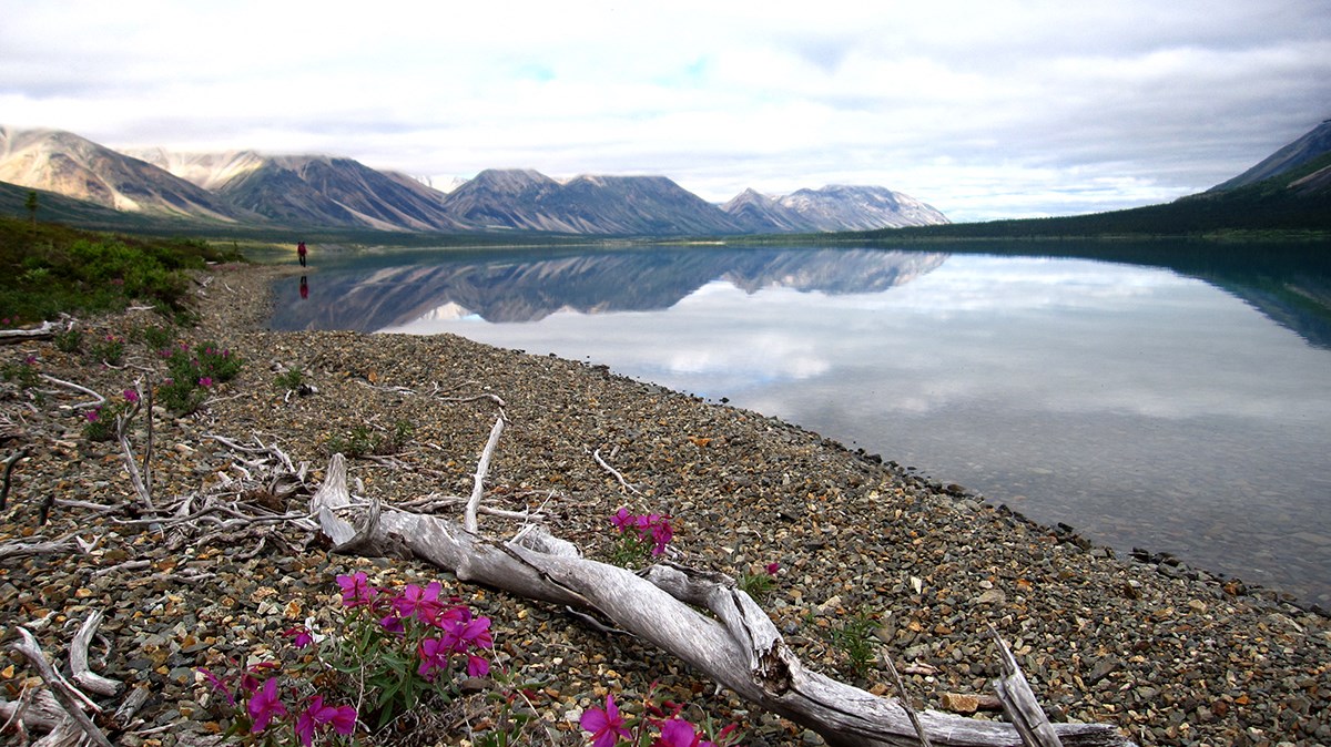 A hiker, mountains, and clouds reflect in a perfectly still a lake.