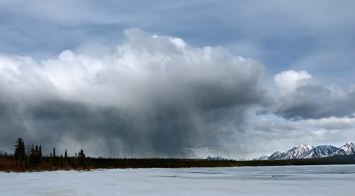 Photo looking across a frozen lake at a storm cloud which is raining in a small area.