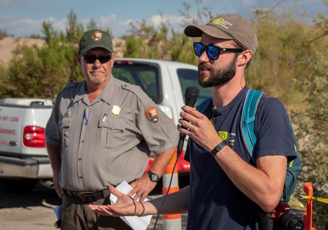 A Park Ranger and a Leave No Trace representative stand together with a microphone to speak about the Spotlight events. There is a truck and desert shrubs behind them.