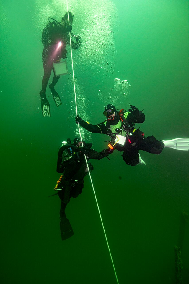 A group of 3 divers use their hands to follow a fixed rope down, releasing clouds of bubbles as they descend.