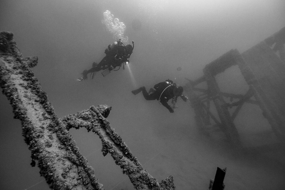 Two divers release cloud of air bubbles as they explore underwater structures in a black and white photo.