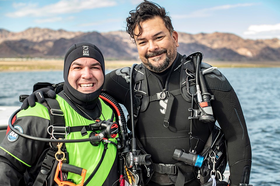 Two divers in wetsuits pose with all their gear prior to dive.