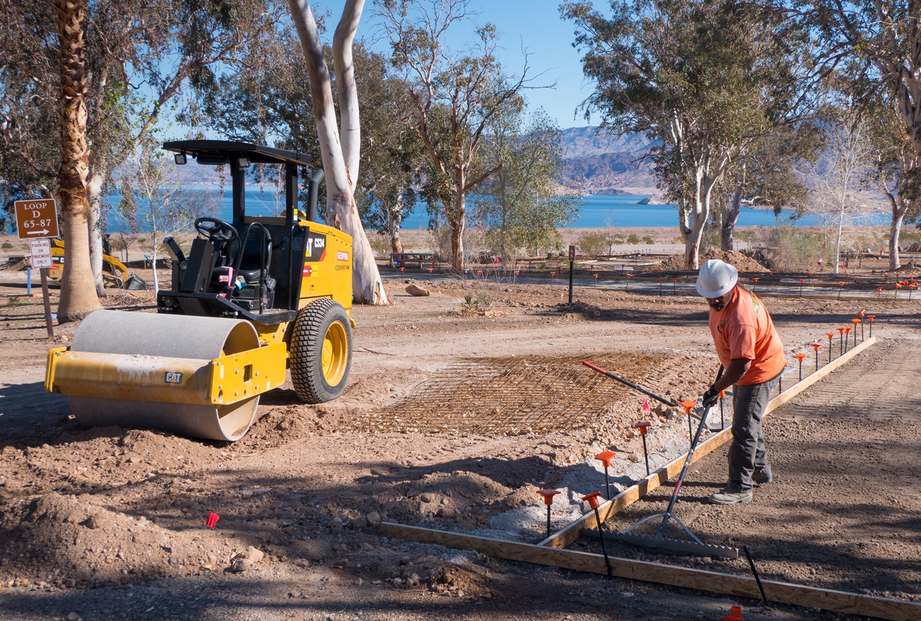 A construction worker with white hard hat and bright orange shirt works with yellow backhoe to improve concrete pad in campground. Lake Mead is in the background.