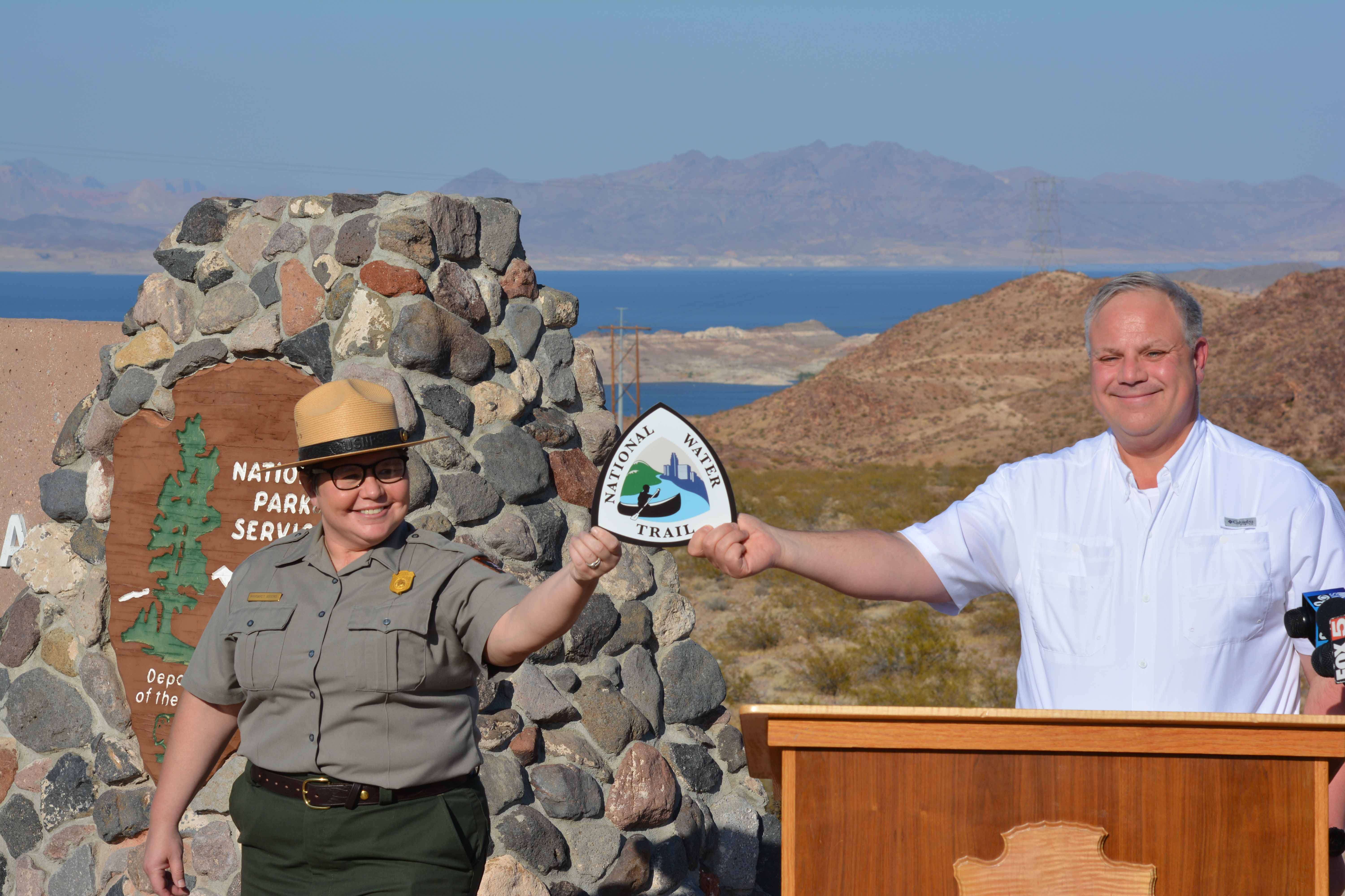 A man and park ranger holding up a sign in front of a Lake Mead Monument.