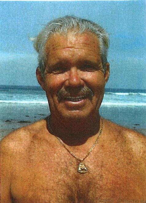 Male with white hair combed back from face stands in front of waves on beach. Man has a gold necklace and is not wearing a shirt. He has a mustache.