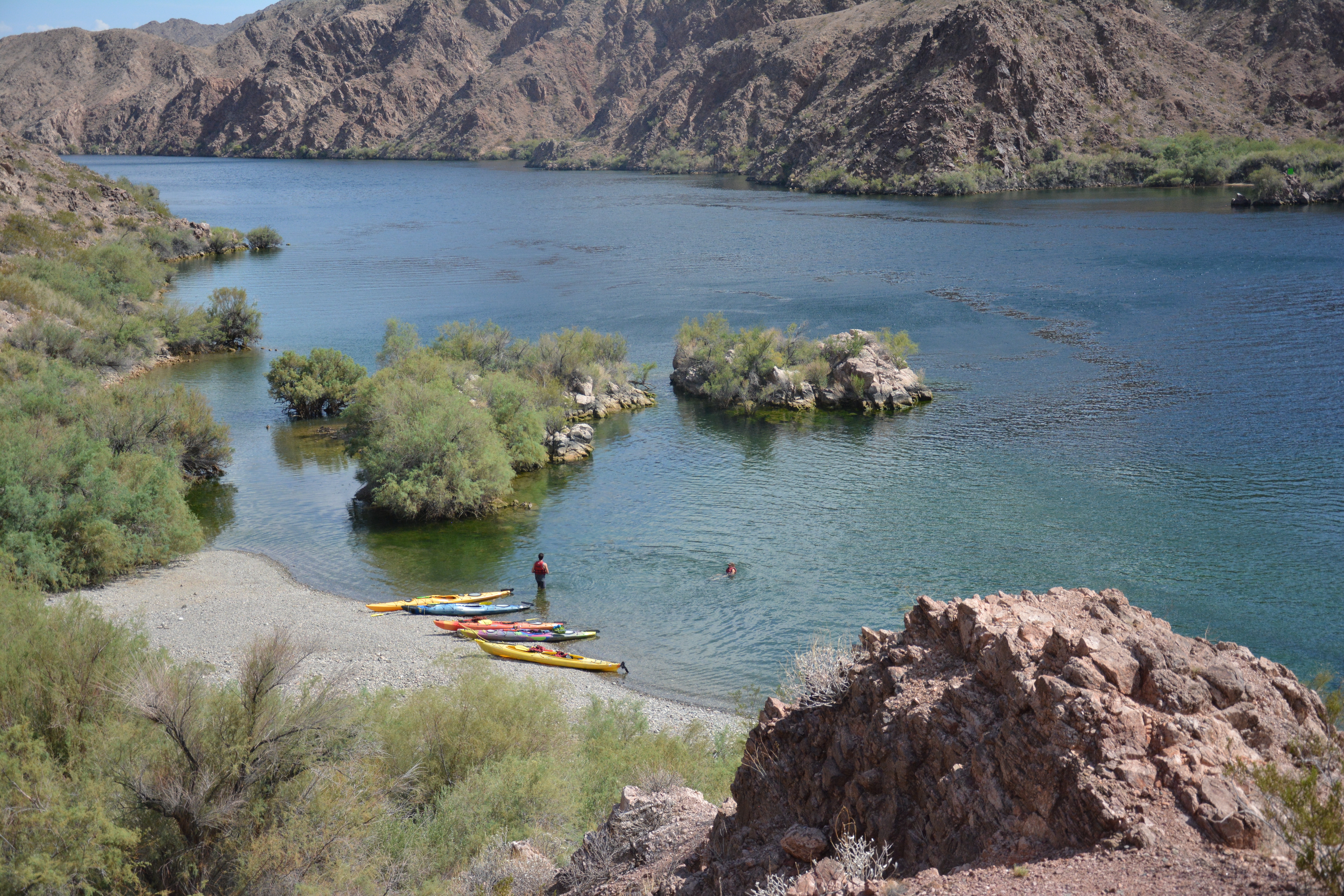 Colorful kayaks are pulled up on a sandy beach in a steep red rock canyon. Two people with life vest swim in the clear cold water.