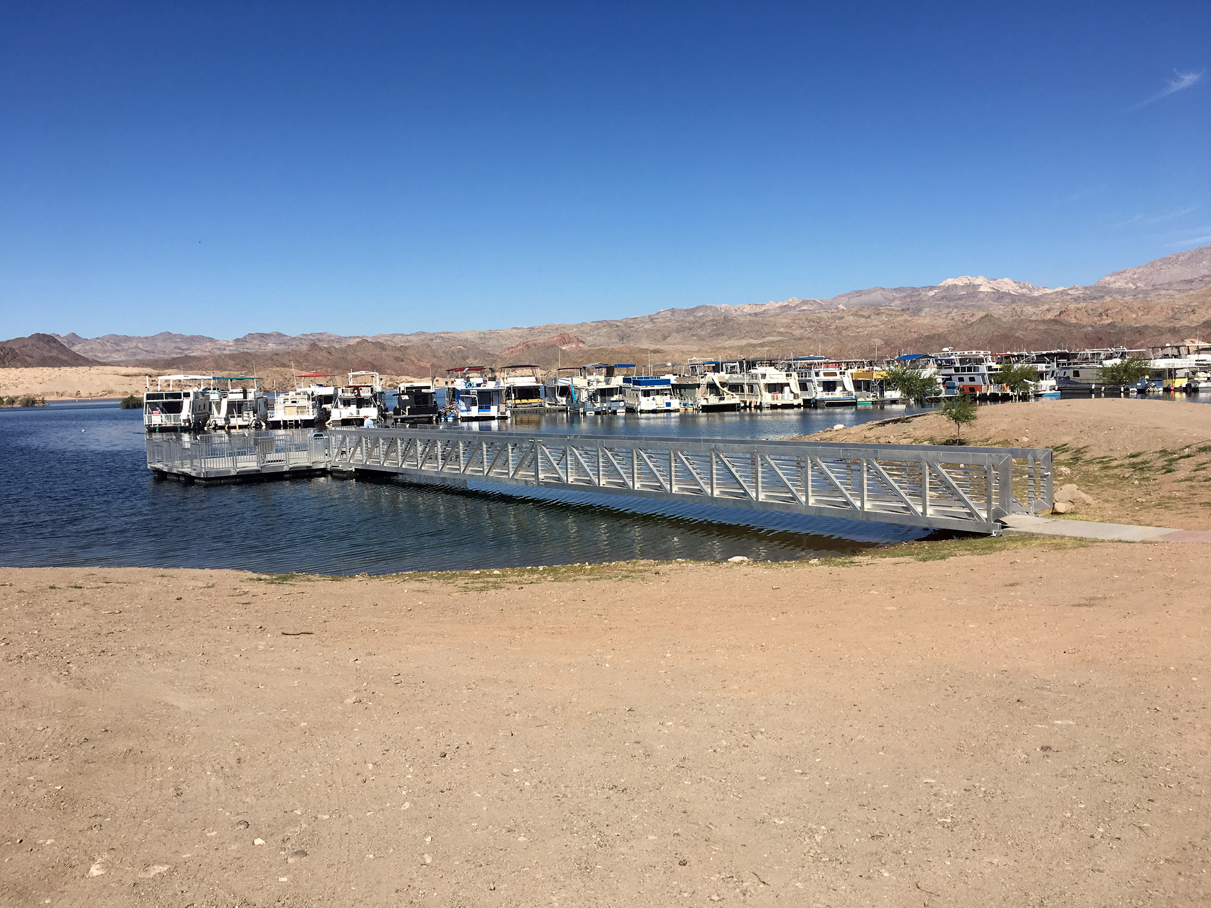 Sandy beach leads to accessible fishing pier. Marina is behind with many houseboats moored. Rocky hills are in the background.