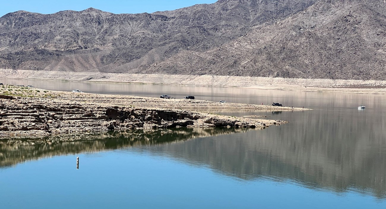 A dirt boat launch ramp is going into the water. Vehicles launching boats are on the ramp. Mountains are in the background.