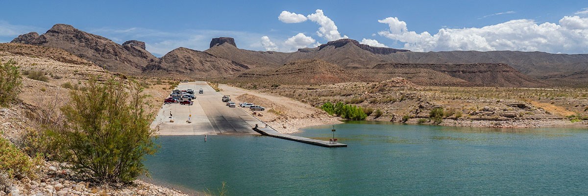 Body of water in foreground, cement launch ramp with vehicles surrounded by desert hill and mountains