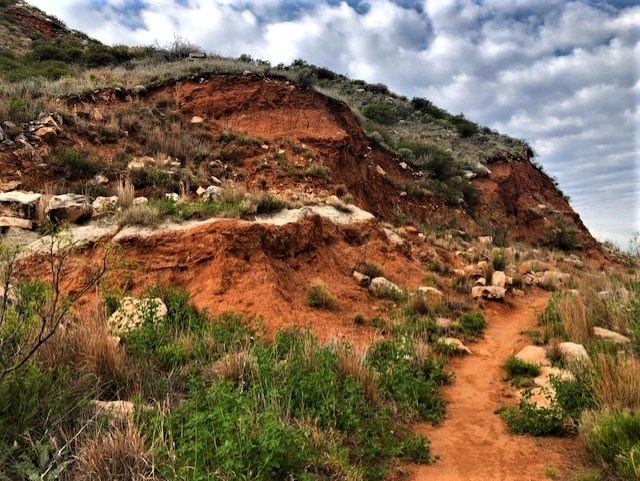 urkey Creek hiking and biking trail in the Permian redbeds.  It is a cloudy day with some blue skies. There is green vegetation along the trail and white dolomite rocks.