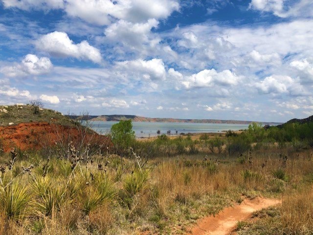 Hiking along Harbor Bay Trail with a view of the blue lake on a cloudy day.  Green vegetation grows along the Permian dirt trail.