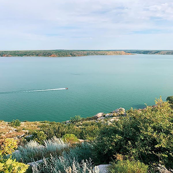 A boater on the lake near Fritch Fortress.  The view is from a rocky mesa with white rocks and green plants.  The lake is teal blue and the sky is blue.