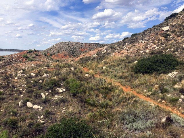 Headed up the trail between the Canadian River Breaks.  The mesas are covered with white rock. The sky is blue and the clouds are puffy white.