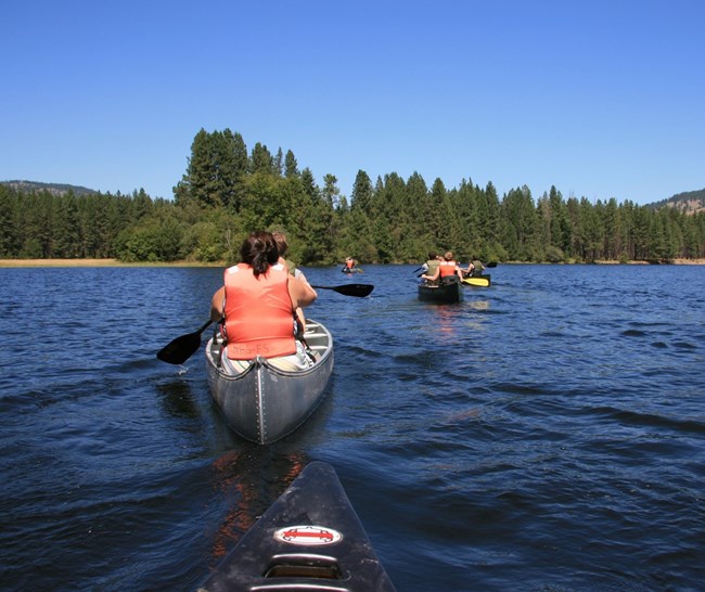 Groups of adults in red life vests paddle on a dark blue lake in silver canoes.