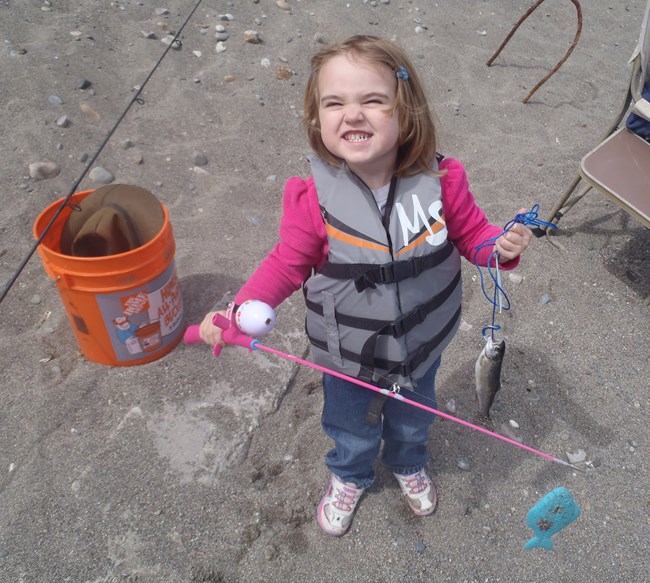 Young blond girl with a big grin holds a pink fishing pole and a small caught fish.