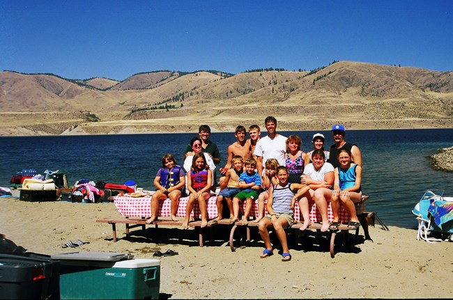 A group of adults and children pose for a photo on the lake shore. The photograph is grainy, as if it was taken a while ago.