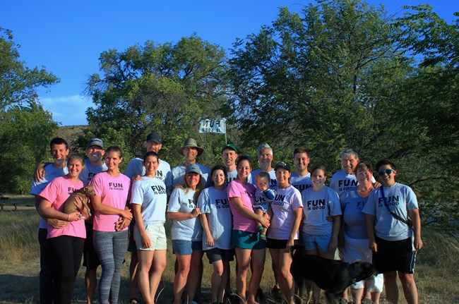 A group of adults, one child, and a dog, pose for a camera with green trees in the background. Each family member is wearing a t-shirt that says "fun is good."