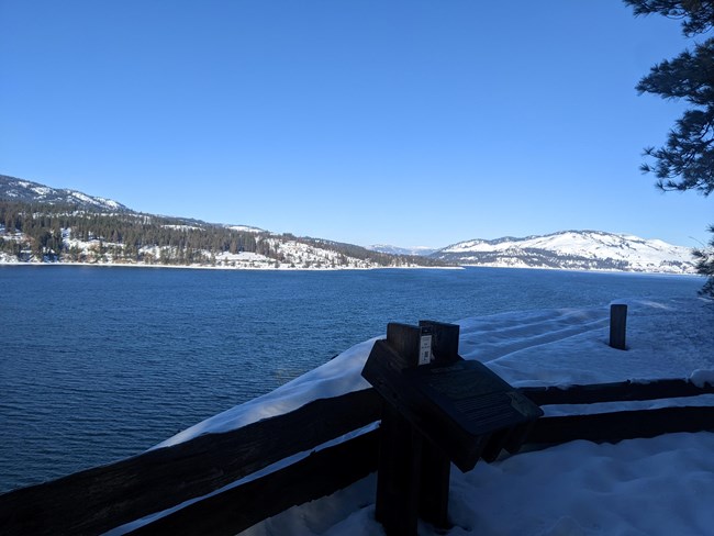 A snowy landscape with short mountains in the background and a blue lake in the foreground.