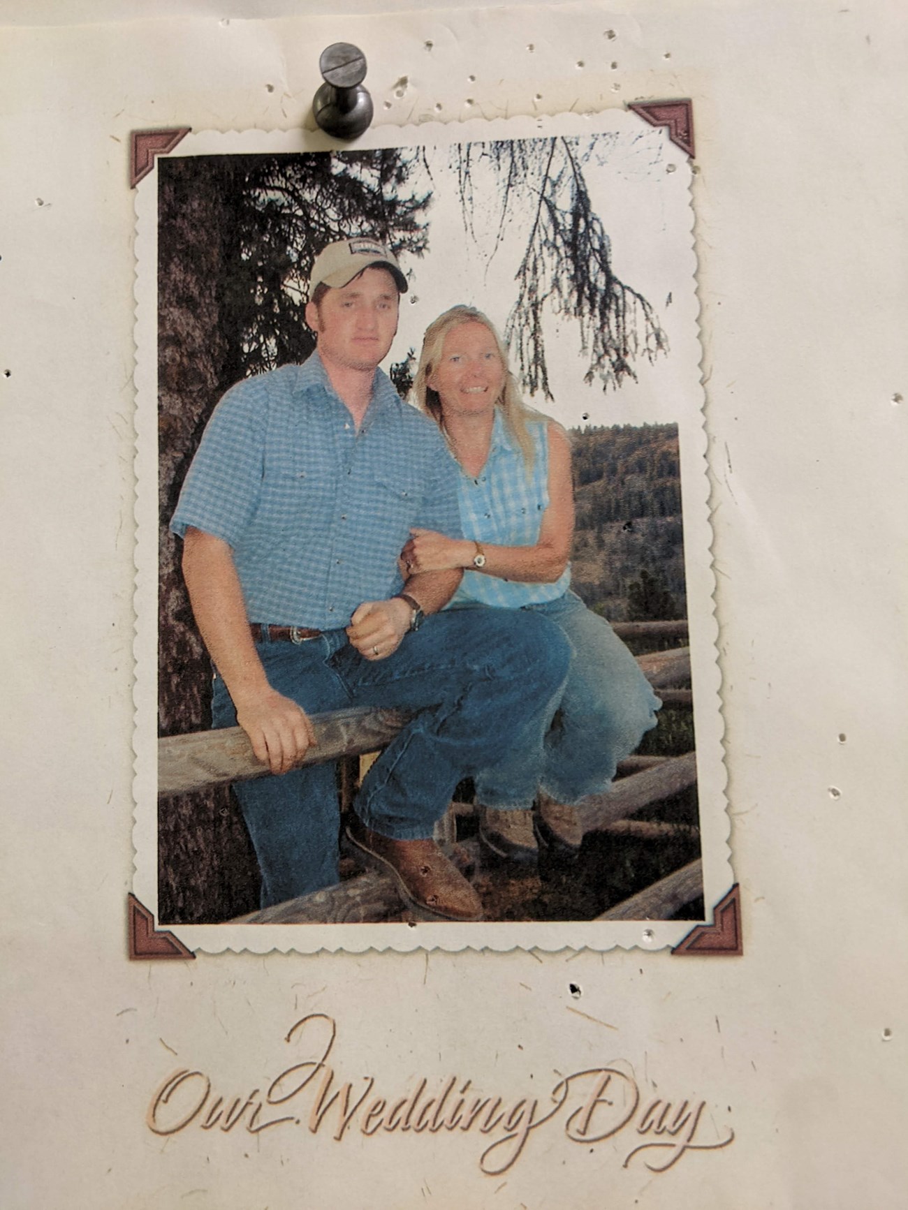 Steve Wedding Photo A photograph is pinned to a cork board: a couple sits on the edge of a fence railing, both dressed in blue plaid, smiling for the camera. Below, it reads "Our Wedding Day."
